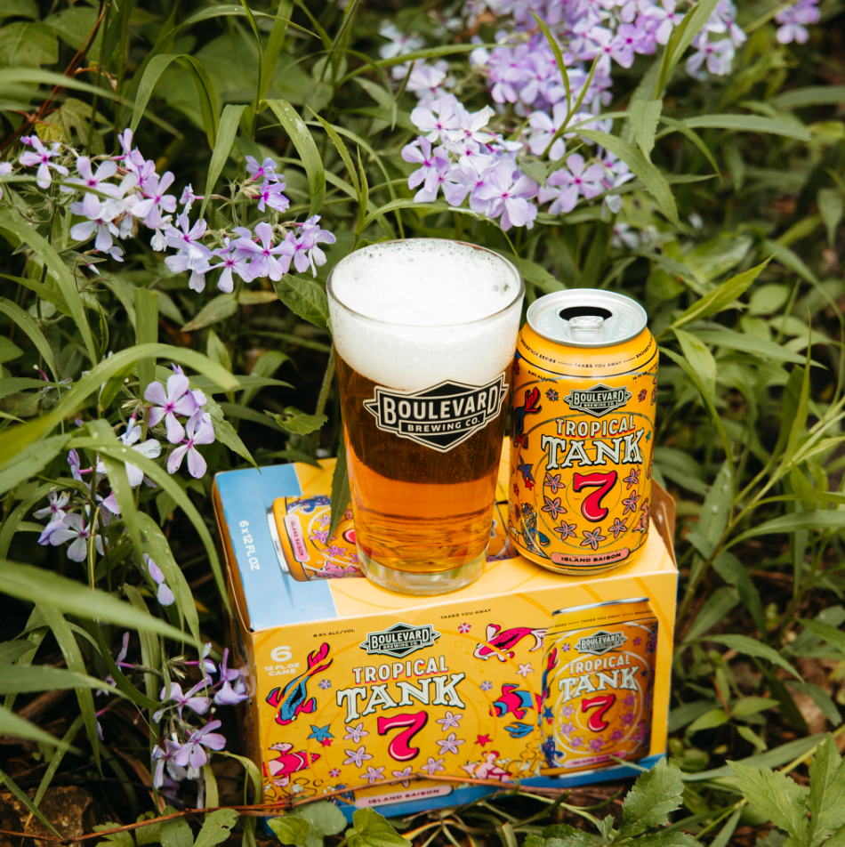 A can and a glass of Boulevard Brewing Co. Tropical Tank 7 beer sit on a six-pack box amid green grass and purple flowers. The glass of golden Tropical Tank 7 gleams with a frothy head, capturing the essence of summer outdoors.